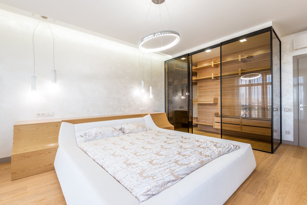 Modern interior design of cozy luxury bedroom with cozy king sized bed and glass wardrobe closet lighted with led chandelier and bedside lamps